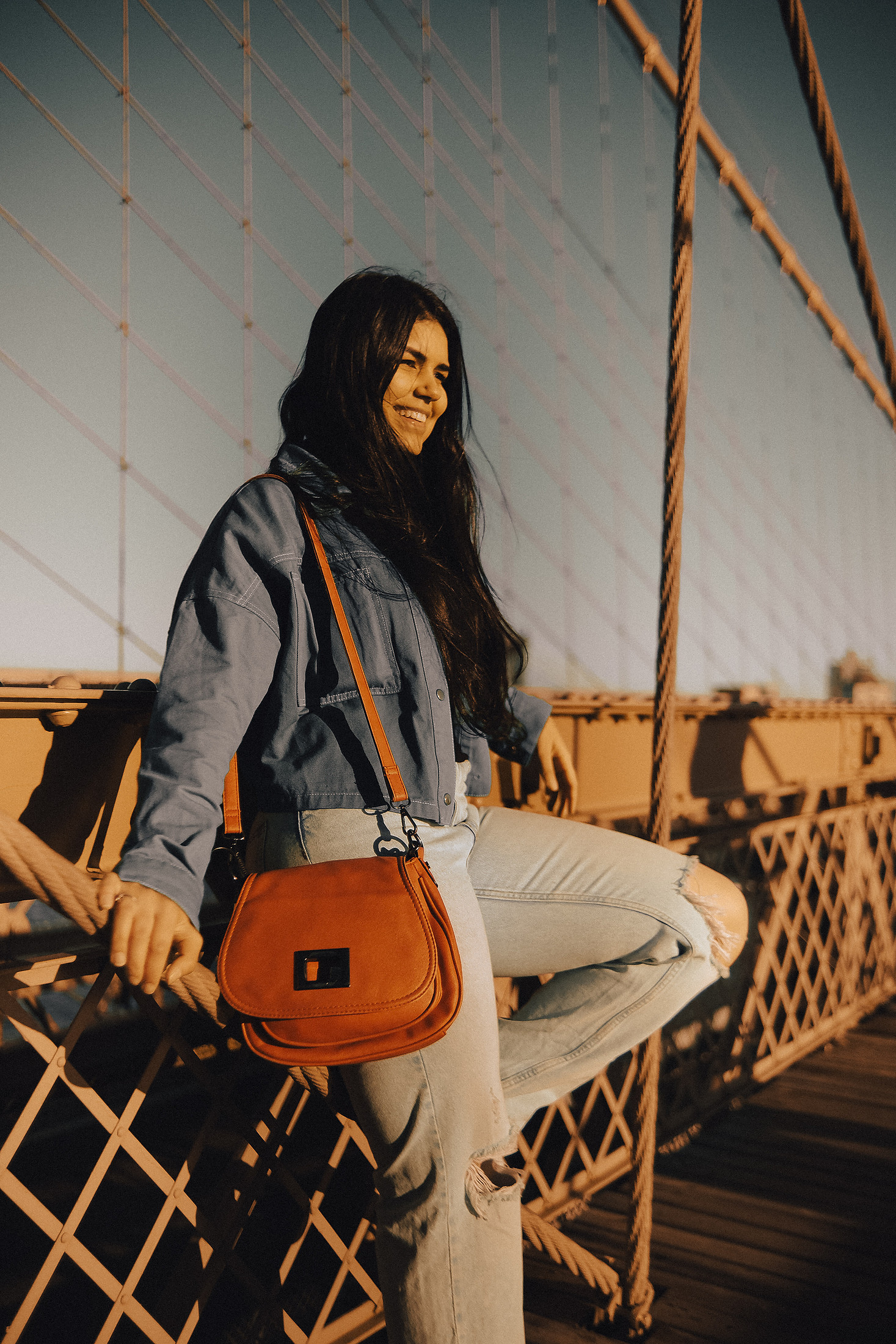Portrait of a woman leaning on the Brooklyn Bridge railing with an orange bag, late afternoon light