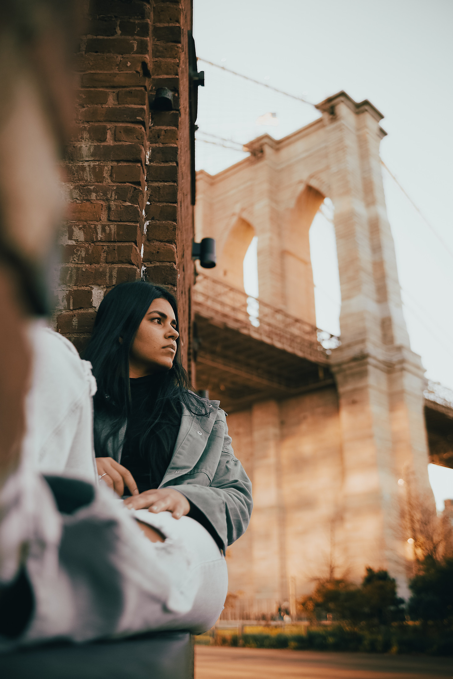 Woman seated against a brick pillar with the Brooklyn Bridge tower behind her at dusk