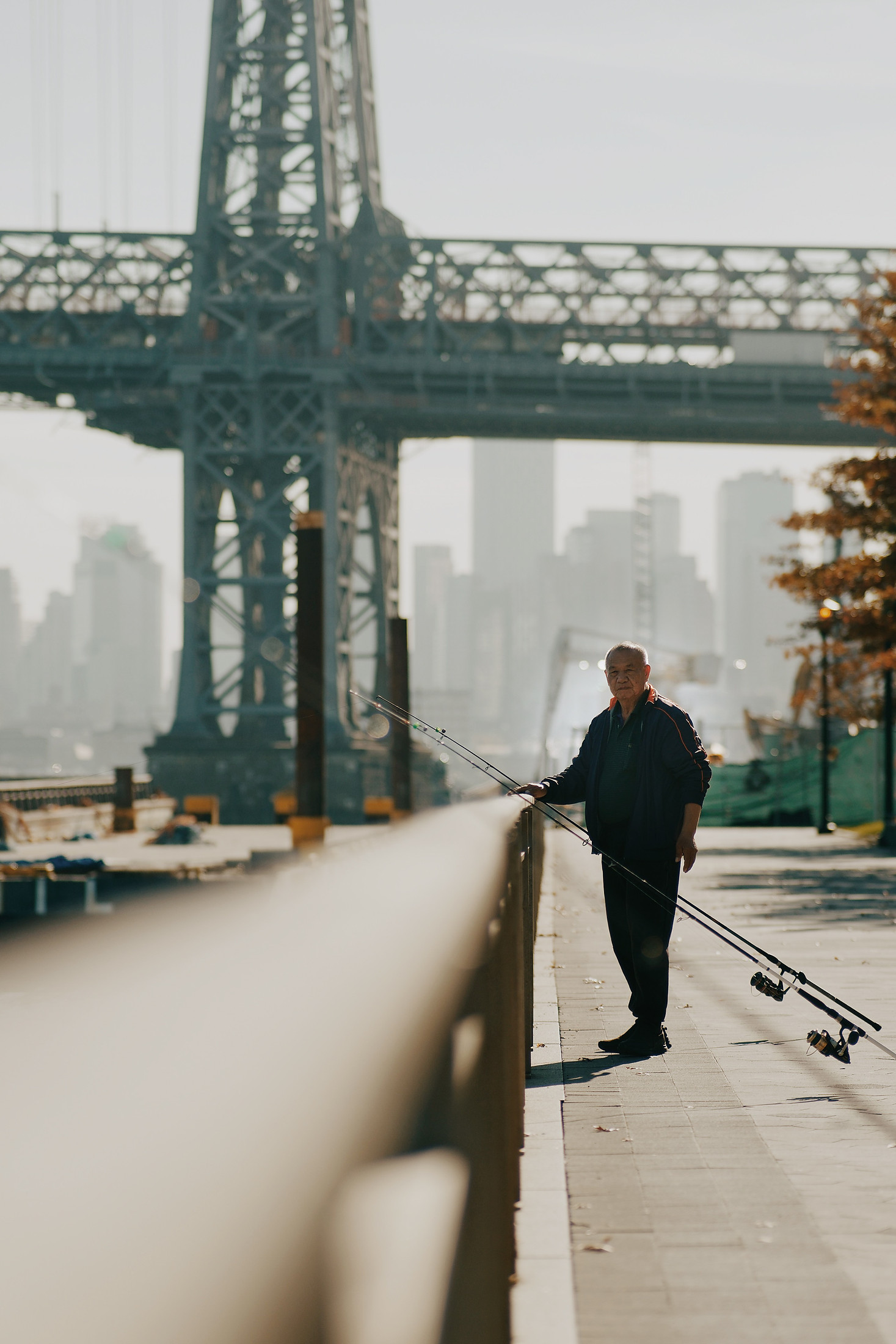 A man carrying fishing rods along the waterfront, with a bridge and the skyline behind him
